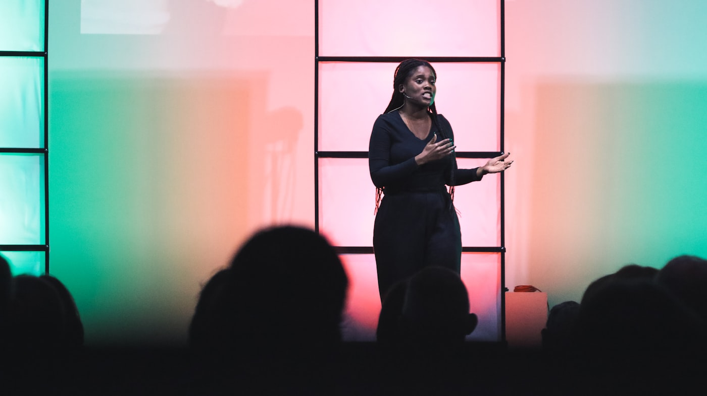 A woman speaking on a conference stage under gradient lights, audience silhouetted in the foreground
